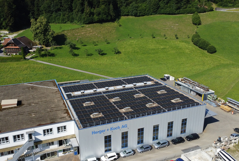Eine Luftaufnahme eines weißen Industriegebäudes mit einem Dach, das mit Solarmodulen bedeckt ist. Auf dem Gebäude steht „Herss + Koch AG“. Um das Gebäude herum sind parkende Autos und im Hintergrund grüne Felder mit Bäumen zu sehen.
