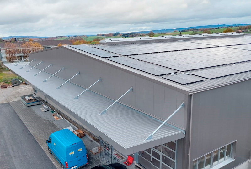 Eine Schrägansicht eines Geschäftsgebäudes mit einem großen, grauen Metalldach, das mit Solarmodulen bedeckt ist. Das Gebäude hat ein Vordach mit Stützstangen, die aus der Fassade herausragen. Neben dem Gebäude parkt ein blauer Lieferwagen. Im Hintergrund sind offene Felder und Bäume zu sehen.