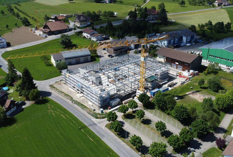 Eine Luftaufnahme einer Baustelle mit einem großen gelben Kran, der über einem Gebäudegerüst thront. Um die Baustelle herum befinden sich Wohnhäuser, grüne Felder und ein paar Bäume, alles in einer ländlichen Landschaft. Verschiedene Baumaterialien sind in der Gegend verstreut.