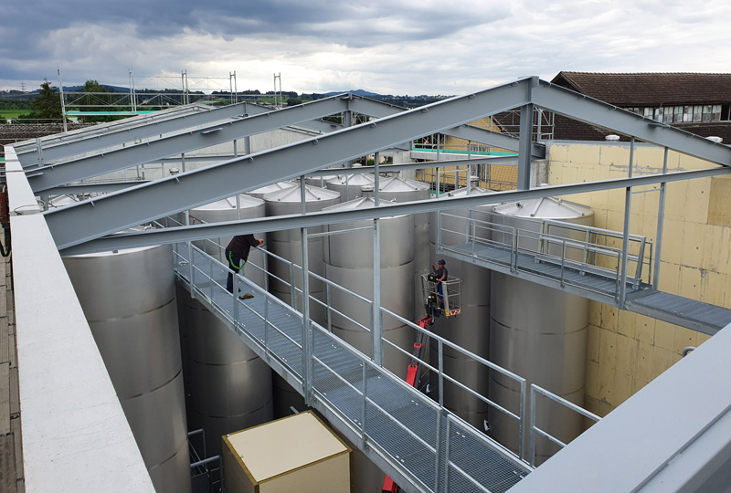 Industrieanlage mit mehreren großen Metalltanks, die durch erhöhte Laufstege miteinander verbunden sind. Arbeiter sind vor Ort; einer bedient einen Aufzug. Die Anlage verfügt über ein teilweise fertiggestelltes Dach mit Balken, die die offenen Flächen einrahmen. Der Himmel ist bedeckt und in der Nähe befinden sich Gebäude.