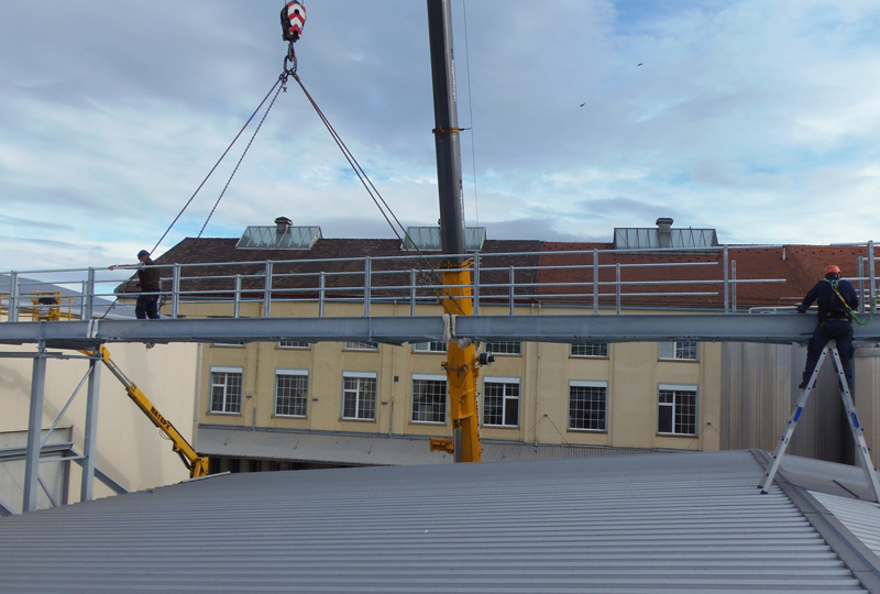 Zwei Bauarbeiter installieren mithilfe eines Krans einen großen Metallträger oder eine brückenartige Struktur. Sie stehen auf beiden Seiten des Trägers, der über einem Gebäude mit mehreren Fenstern thront. Der Himmel ist teilweise bewölkt.