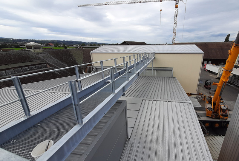 Blick auf eine Baustelle mit einem neu errichteten erhöhten Gehweg mit Geländern, der zwei Gebäude mit Metalldächern verbindet. Kräne im Hintergrund unterstützen weitere Bauarbeiten. Der Himmel ist bedeckt und das Gebiet zeigt eine laufende Entwicklung.