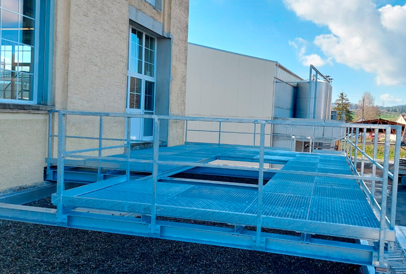 An der Seite eines Beiss-Industriegebäudes ist eine Metallgitterplattform mit Sicherheitsgeländer angebracht. Die Plattform erstreckt sich nach außen und überblickt einen mit Kies bedeckten Boden. Im Hintergrund sind weitere Industriegebäude und ein teilweise bewölkter Himmel zu sehen.