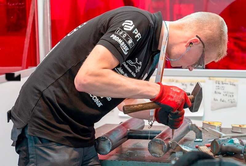 Eine Person in schwarzem T-Shirt und roten Handschuhen bearbeitet mit Hammer und Meißel Metallrohre auf einer Werkbank. Die Person trägt außerdem eine Schutzbrille und einen Gehörschutz. Im Hintergrund sind ein leuchtend roter Bildschirm und einige an die Tafel gepinnte Notizen zu sehen.