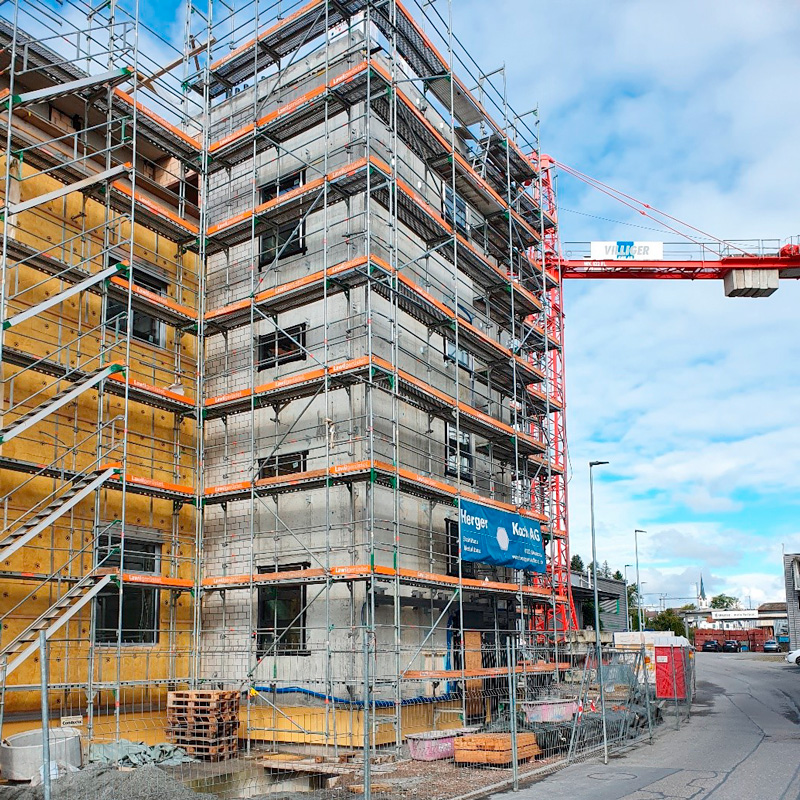 Eine Baustelle, auf der ein mehrstöckiges Gebäude im Bau ist. Ein Gerüst umgibt das Gebäude, Baumaterialien und Maschinen sind sichtbar. Im Hintergrund ragt ein Kran vor einem teilweise bewölkten Himmel auf.