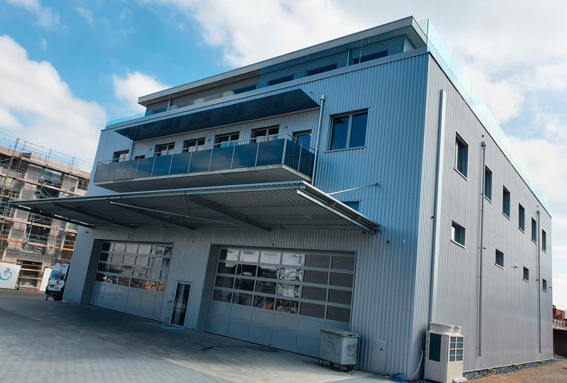 Ein modernes, mehrstöckiges Industriegebäude mit einer grauen Metallfassade und großen Glasgaragentoren im Erdgeschoss. Aus dem zweiten Stock ragt ein kleiner Balkon heraus, und auf der linken Seite des Bildes ist eine Baustelle zu sehen. Der Himmel ist teilweise bewölkt.