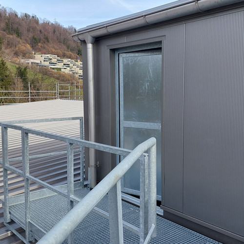 Eine graue Metalltür mit Milchglas in einem Wellblechgebäude ist über einen Metallsteg mit Geländer zugänglich. Im Hintergrund sind Bäume und ein Hügel mit modernen Gebäuden zu sehen. Der Himmel ist klar und blau.