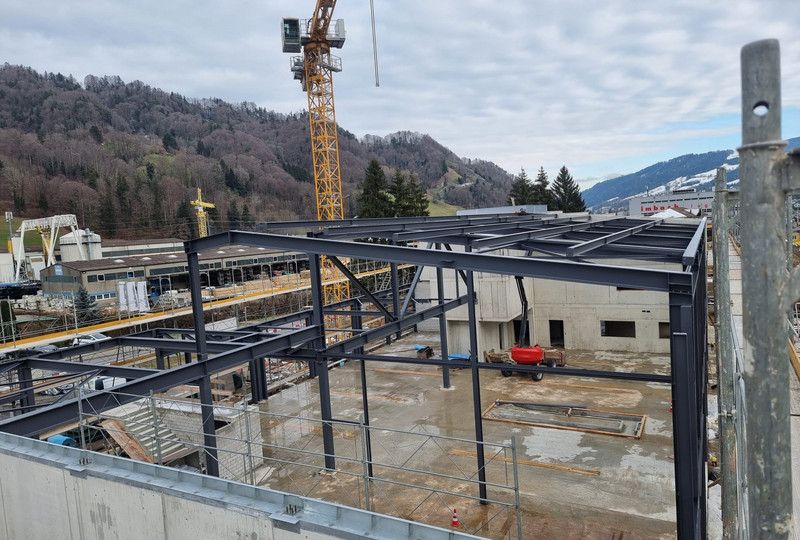 Eine Baustelle mit dem Metallgerüst eines Gebäudes im Bau. In der Mitte links steht ein Kran, im Hintergrund sind schneebedeckte Berge zu sehen. In der Umgebung der Baustelle befinden sich Industriegebäude und Bäume. Der Himmel ist bewölkt.