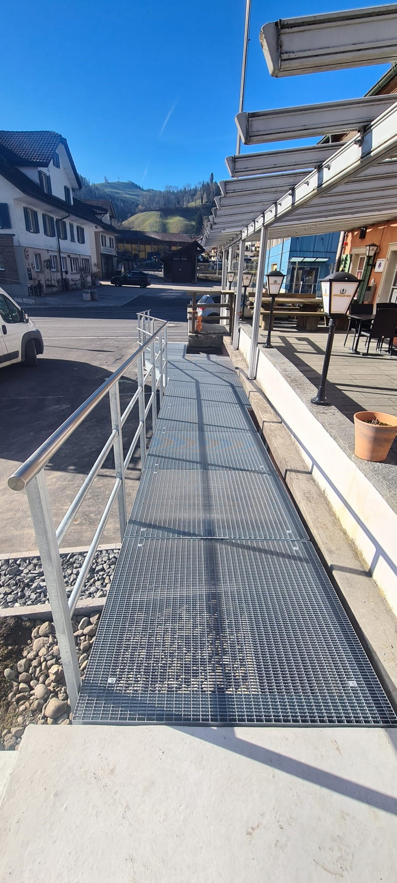 Eine Metallrampe mit Handläufen auf beiden Seiten führt zu einem Gebäudeeingang und bietet behinderten Menschen Zugang. Die Rampe befindet sich in einem sonnigen Außenbereich, umgeben von Gebäuden, einem geparkten weißen Lieferwagen und einem Pflanzkübel mit Erde.