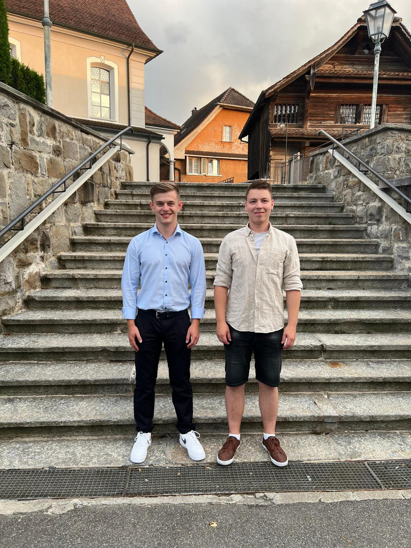 Zwei junge Männer stehen nebeneinander auf einer Steintreppe im Freien und lächeln beide in die Kamera. Einer trägt ein hellblaues Hemd mit schwarzer Hose und weißen Turnschuhen, während der andere ein beiges Hemd mit schwarzen Shorts und braunen Schuhen trägt. Im Hintergrund sind mehrere Gebäude zu sehen.