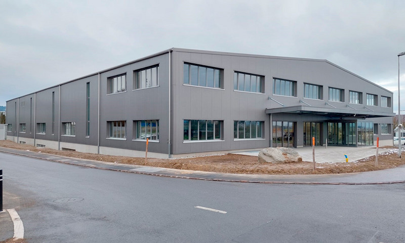 Gezeigt wird ein großes Industriegebäude mit grauer Außenfassade und mehreren Fenstern. Das Gebäude hat ein Flachdach und einen überdachten Eingang mit Glastüren. Die umgebende Landschaft umfasst eine gepflasterte Straße, kahlen Boden und ein paar kleine Pfosten und Felsen. Der Himmel ist bedeckt.