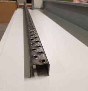 Ein langer U-förmiger Metallkanal mit regelmäßig angeordneten kreisförmigen Löchern auf einer Seite liegt auf einer flachen, hellen Oberfläche. Oberfläche und Hintergrund enthalten verschiedene Objekte und Strukturen, die leicht unscharf sind.