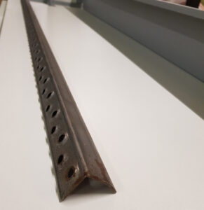 Eine Nahaufnahme eines verrosteten Winkeleisens aus Metall mit Löchern, das auf einer weißen Oberfläche liegt. Das Winkeleisen verläuft diagonal von der unteren linken Ecke des Bildes zur oberen rechten Ecke, wobei der Rost und die Löcher deutlich sichtbar sind.